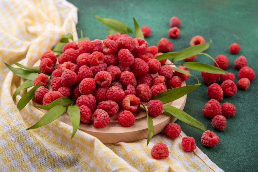 side-view-of-raspberries-with-leaves-on-cutting-board-on-plaid-cloth-and-on-green.jpg