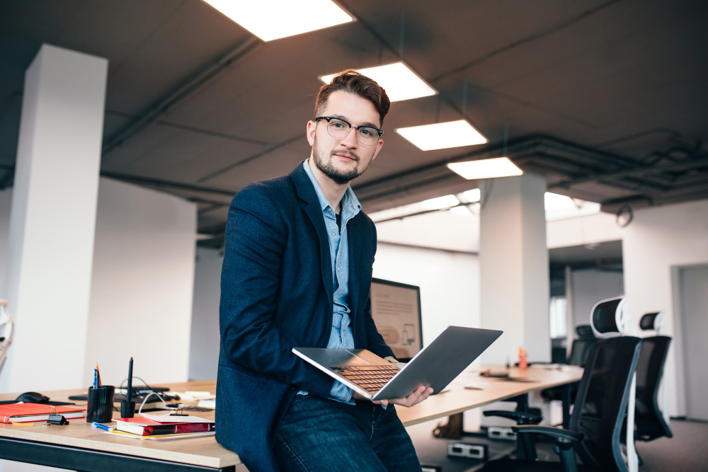 attractive-man-in-glassess-is-sitting-near-the-workplace-in-office-he-wears-blue-shirt-dark-jacket-he-holds-laptop-and-looks-to-the-camera.jpg